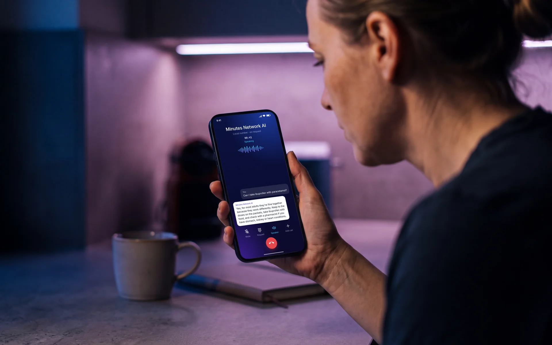 A woman in her kitchen in the evening talking to Minutes Network AI on speakerphone, the call screen showing her question and the spoken answer