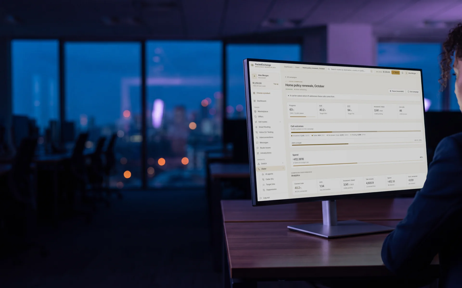 A contact-centre lead at dusk watching a running PacketExchange dialer campaign on a monitor, with progress, answer rate, call length, live calls and spend against budget.