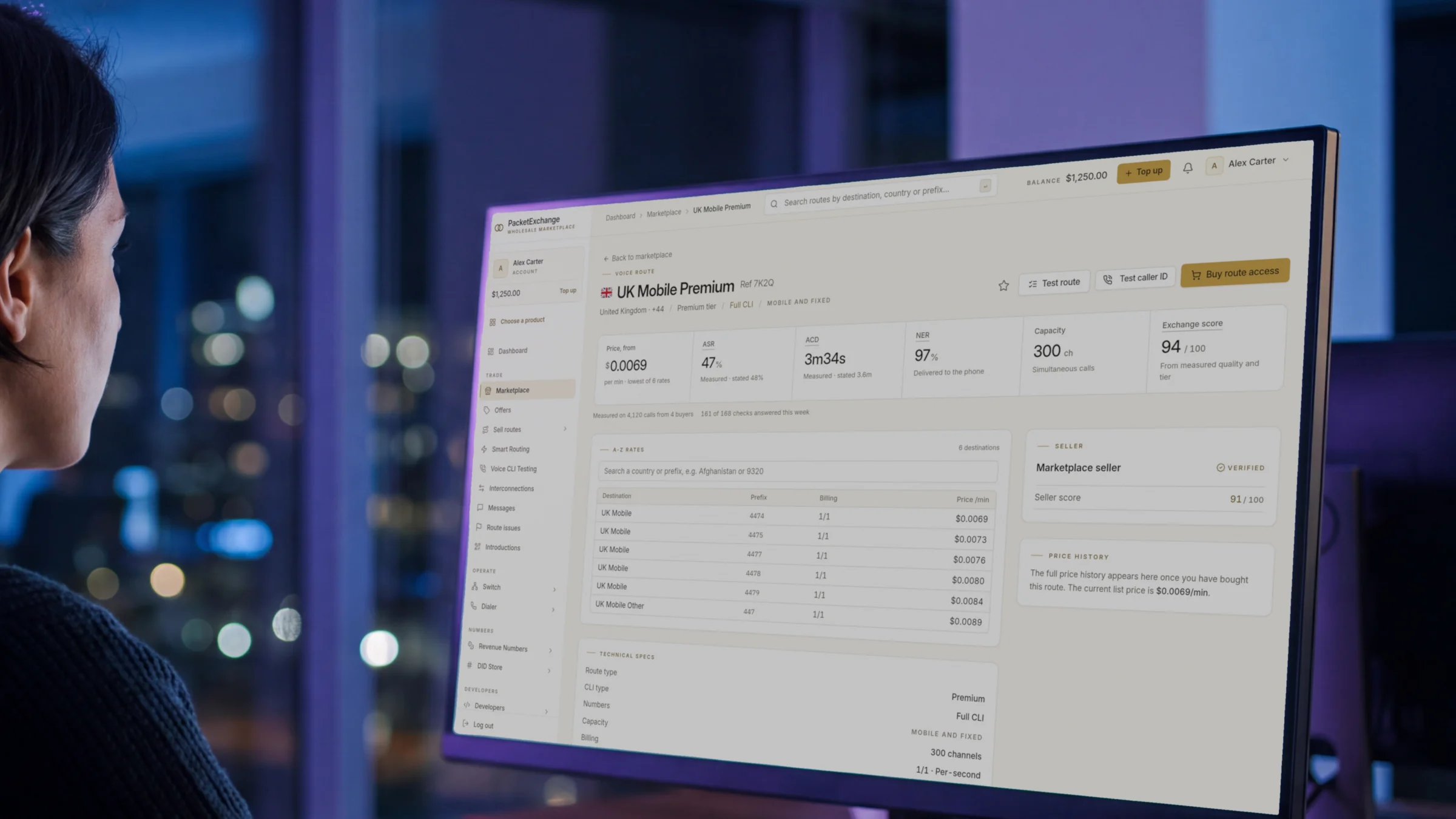 A buyer at a large monitor at night studying a PacketExchange route page for UK Mobile Premium, with price, measured ASR, ACD, NER, capacity and Full CLI