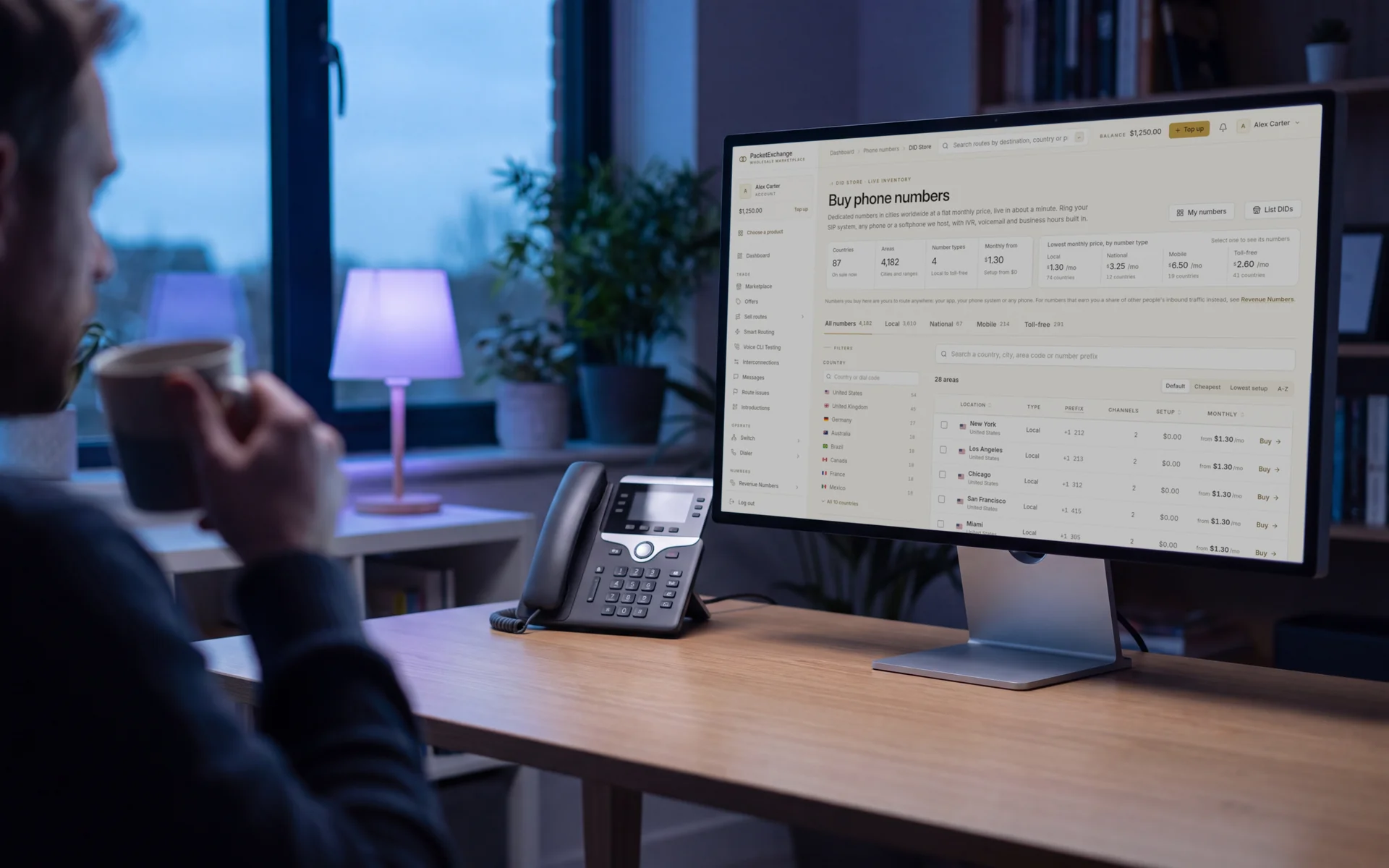 A small office at dusk with a desk phone beside a monitor showing the PacketExchange number store: countries, areas, number types and local numbers by city with monthly prices