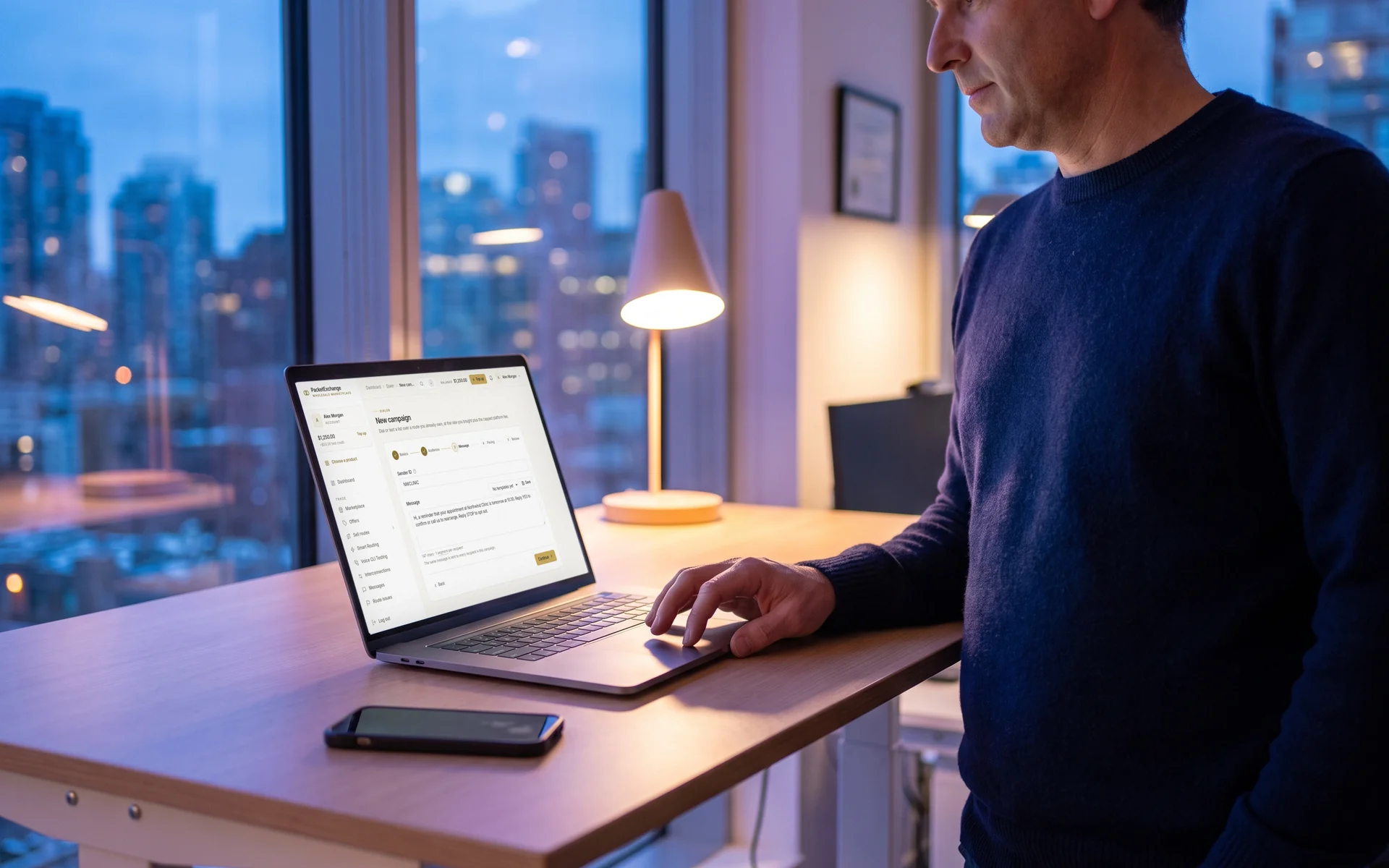 A clinic operations manager at a standing desk with a laptop showing a PacketExchange SMS campaign being written, with the sender ID and an appointment reminder message
